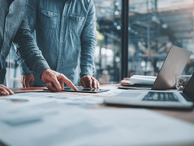 Close-up of two professionals in denim shirts leaning over a desk covered with blueprints and documents, analyzing details on a tablet beside a laptop — How to Deal with Project Cost Overruns article