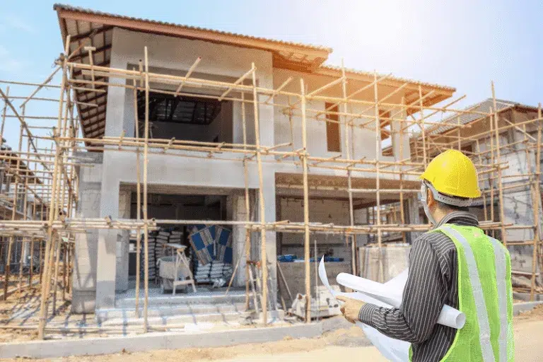 A construction supervisor in a safety vest and hard hat reviewing building plans on-site, inspecting the progress of a two-story home under active construction — 10 Mistakes to Avoid When Building a Home article