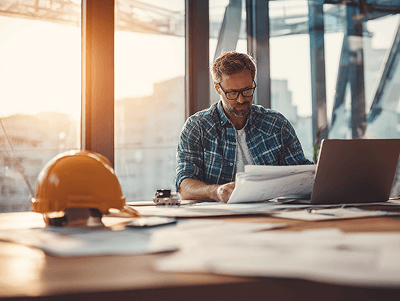 A construction project manager reviews architectural blueprints at his desk during sunset, with a laptop and safety helmet nearby in a modern office overlooking an active building site - How to Start Your Own Construction Business article