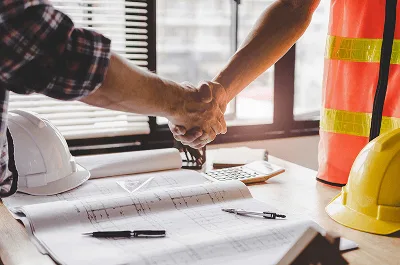 10 Tips for Completing Construction Projects On Time and On Budget 3 Two people shake hands over construction plans on a desk, symbolizing agreement or partnership — 10 Tips for Completing Construction Projects On Time and On Budget article