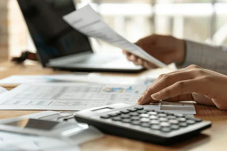 Construction Contingency: All You Need to Know 2 Close-up of a construction cost manager reviewing budget documents and using a calculator beside a laptop — Construction Contingency All You Need to Know article