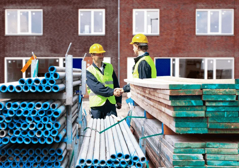 Blog 10 Two construction workers in safety gear shake hands on-site between stacks of building materials — Types of Subcontractors in Construction article