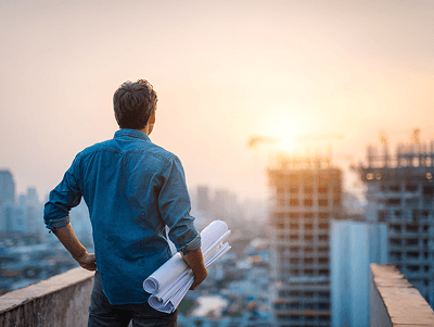 A man stands on a rooftop at sunset, holding blueprints and overseeing high-rise developments — The Role of Construction Quantity Surveyors article
